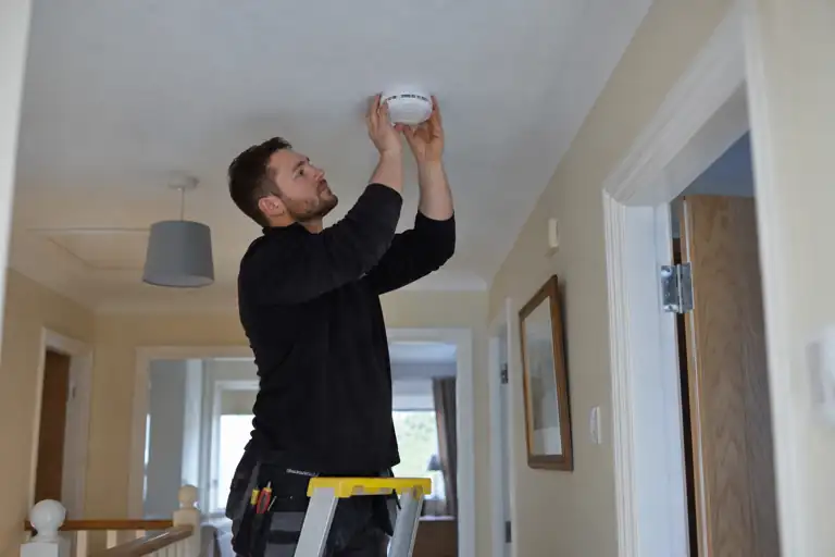 Electrician fitting a smoke alarm to a domestic hallway ceiling.